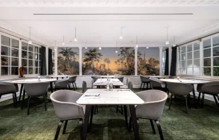 en::Room with square and round tables, grey and black chairs, a wallpapered back wall, and windows opening onto living areas on the left and the inner courtyard on the right. fr::Salle avec tables carrées et rondes, chaises grises et noires, mur du fond tapissé, fenêtres ouvertes sur les espaces de vie à gauche et sur la cour intérieure à droite.