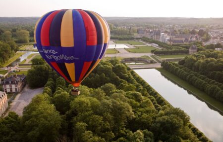 en::Aerial view of the Château de Fontainebleau, its French formal gardens, and the surrounding forest. fr::Vue aérienne du château de Fontainebleau, de ses jardins à la française et de la forêt environnante, avec au premier plan une montgolfière et des personnes dans la nacelle.