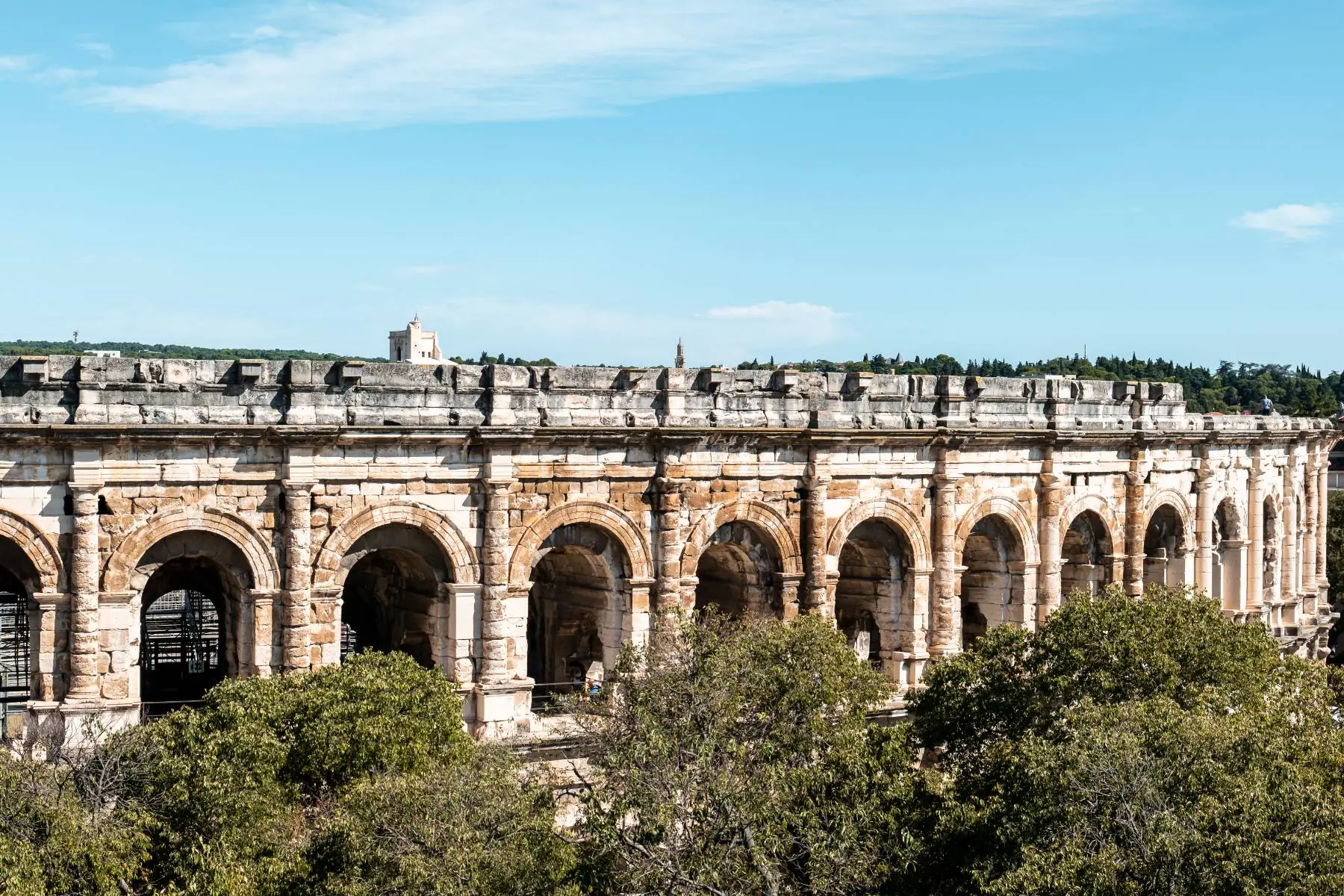 en:: View of the Nîmes Arena photographed from above through surrounding vegetation, with other monuments visible in the distance. fr:: Les arènes de Nîmes photographiées en hauteur depuis la végétation environnante, avec d’autres monuments visibles au loin.