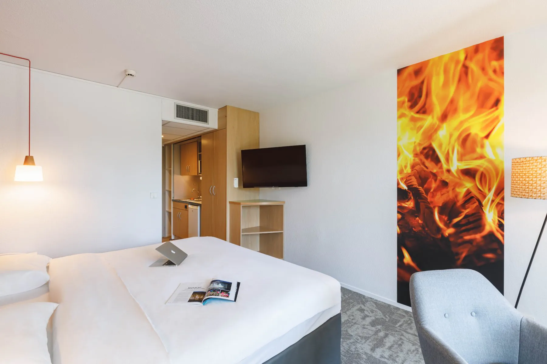 en::Double bed with magazine and laptop on the bed, facing a wall-mounted TV, corner wooden shelving unit, wall artwork depicting fire, grey chair in the corner. fr::Lit double avec magazine et ordinateur sur le lit, face à une TV murale, meuble d’angle en bois avec étagères, photographie murale représentant un feu, chaise grise dans l’angle
