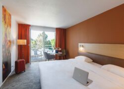 en::Double bed with wooden and leather headboard, lights on, wall section with flame-pattern wallpaper, room in brown and beige tones opening onto a furnished balcony. fr::Lit double avec tête de lit en bois et cuir, luminaires allumés, pan de mur avec tapisserie à motifs de flammes, chambre aux tons marron et beige ouverte sur un balcon aménagé.