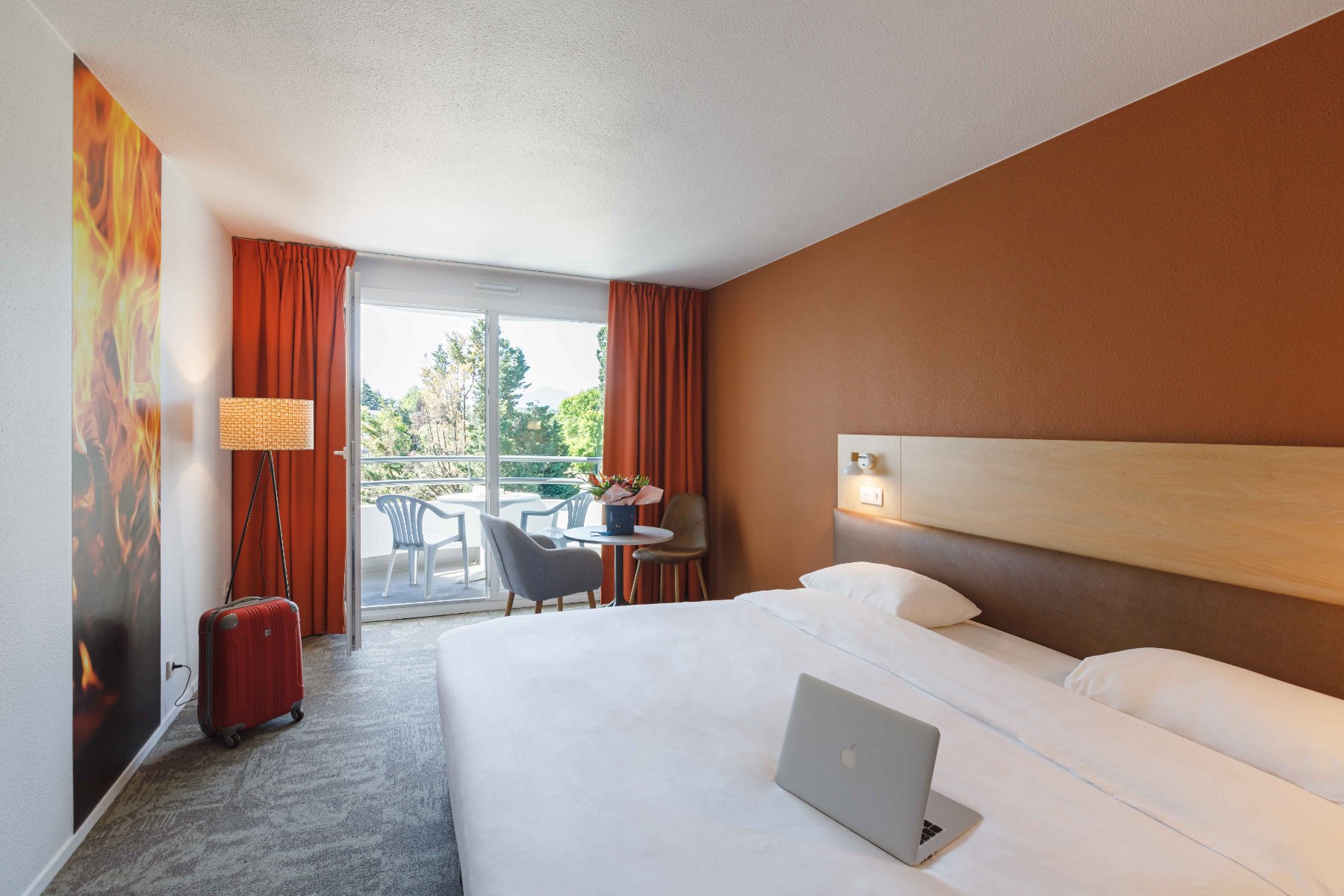 en::Double bed with wooden and leather headboard, lights on, wall section with flame-pattern wallpaper, room in brown and beige tones opening onto a furnished balcony. fr::Lit double avec tête de lit en bois et cuir, luminaires allumés, pan de mur avec tapisserie à motifs de flammes, chambre aux tons marron et beige ouverte sur un balcon aménagé.