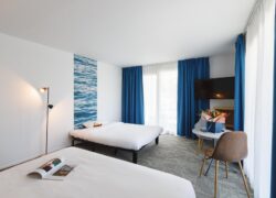 en::Double bed and extra bed against a wall with wave-pattern wallpaper, blue curtains, table with flowers. fr::Lit double et lit d’appoint contre un mur à motifs vagues, rideaux bleus, table avec bouquet de fleurs.