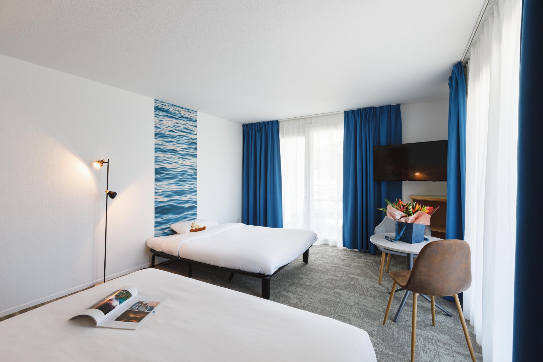 en::Double bed and extra bed against a wall with wave-pattern wallpaper, blue curtains, table with flowers. fr::Lit double et lit d’appoint contre un mur à motifs vagues, rideaux bleus, table avec bouquet de fleurs.