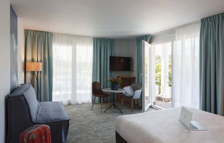 en::Open glass door to the park and balcony, blue sofa bed with red suitcase, double bed with laptop. fr::Porte-fenêtre ouverte sur le parc et le balcon, canapé-lit bleu avec valise rouge, lit double avec ordinateur.