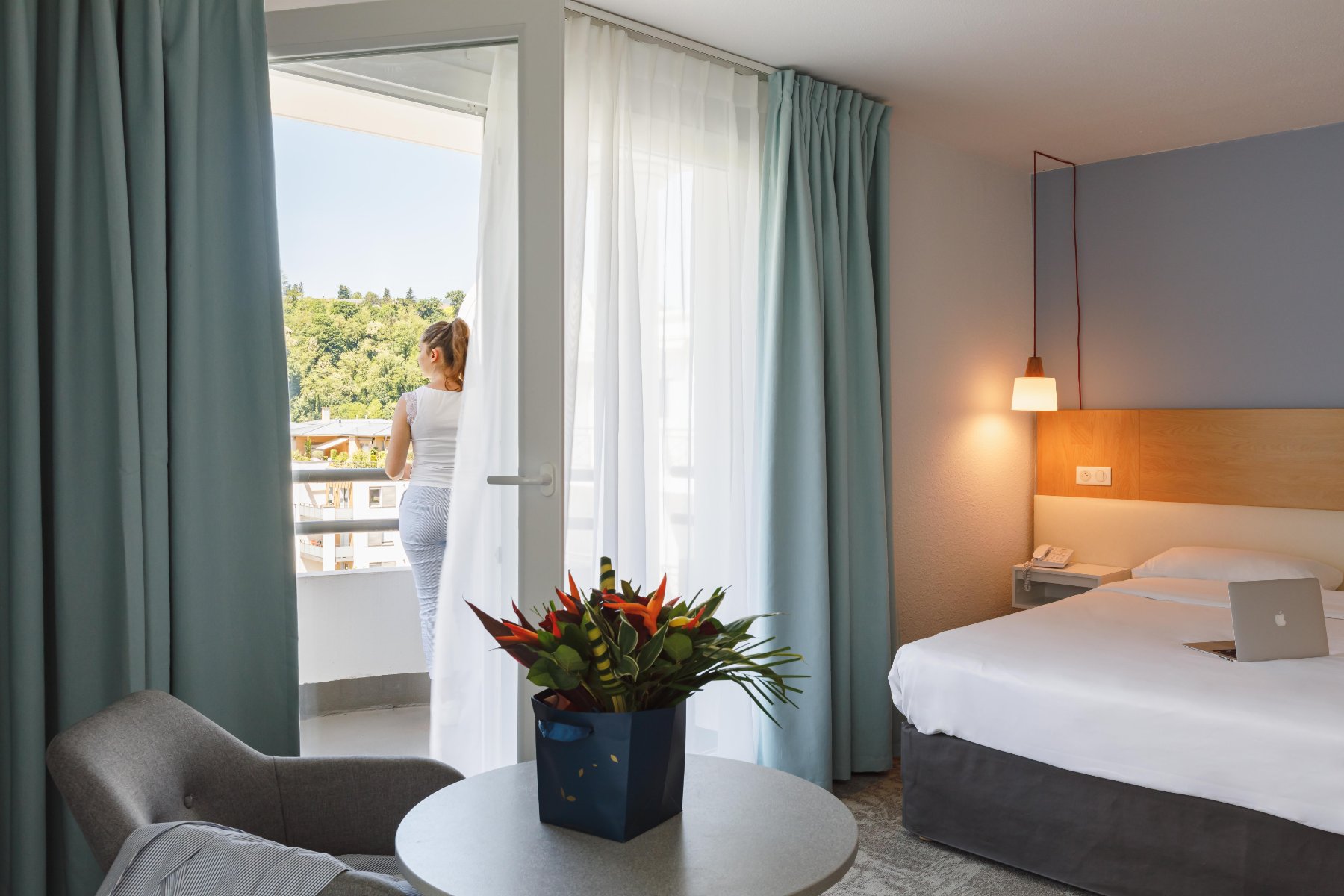 en::Woman seen from behind on a balcony overlooking greenery, jacket on a chair, laptop on the bed, bedside light on. fr::Femme de dos au balcon avec vue sur la végétation, veste sur un fauteuil, ordinateur sur le lit, lampe allumée.