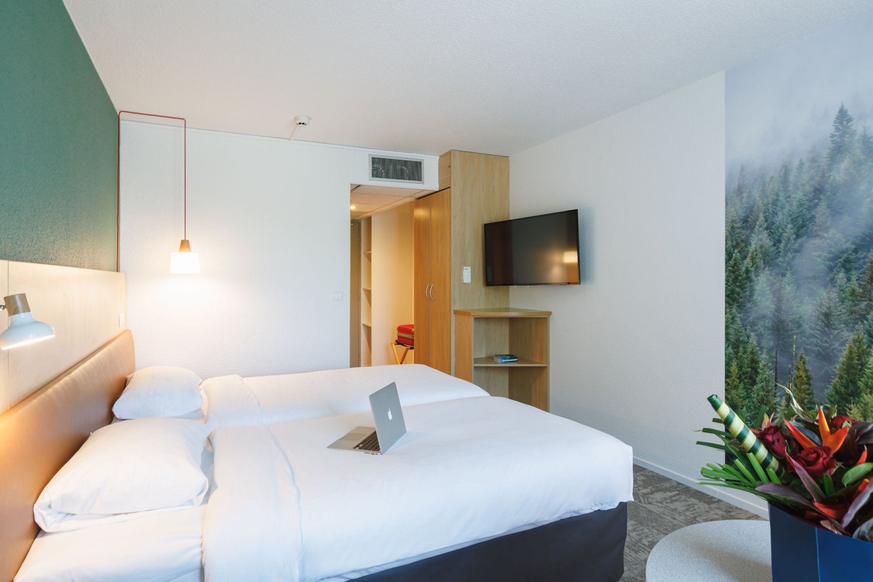 en::Twin beds with wooden and leather headboards, large wall photo of a forest, wall-mounted TV, bouquet of flowers on the bedside table. fr::Lits twin avec têtes de lit bois et cuir, grande photographie de forêt sur le mur, télévision murale, bouquet de fleurs sur la table près du lit.