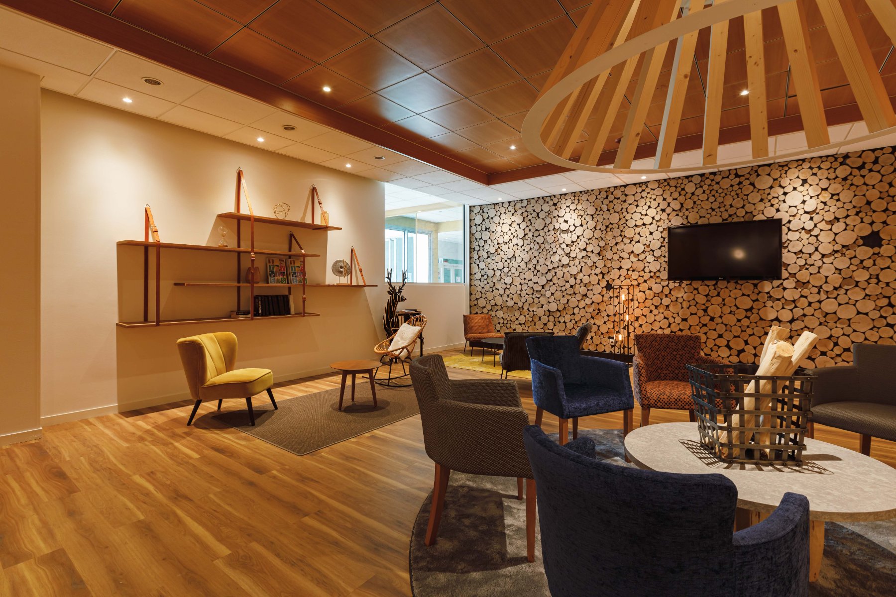 en::Room with wooden floor, wall with round wood patterns, wooden chandelier, shelves with straps, table center with logs and deer décor. fr::Salle avec parquet bois, mur en rondins de bois, lustre bois, étagères suspendues, centre de table avec bûches et décor cerf.