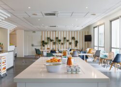 en::White central island with breakfast setup, room with round and square tables, colorful chairs, wall with hanging plants and corner TV. fr::Îlot central blanc avec petit-déjeuner, salle avec tables rondes et carrées blanches, chaises multicolores, mur végétalisé et TV en angle.