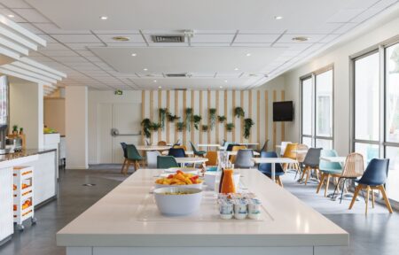en::White central island with breakfast setup, room with round and square tables, colorful chairs, wall with hanging plants and corner TV. fr::Îlot central blanc avec petit-déjeuner, salle avec tables rondes et carrées blanches, chaises multicolores, mur végétalisé et TV en angle.