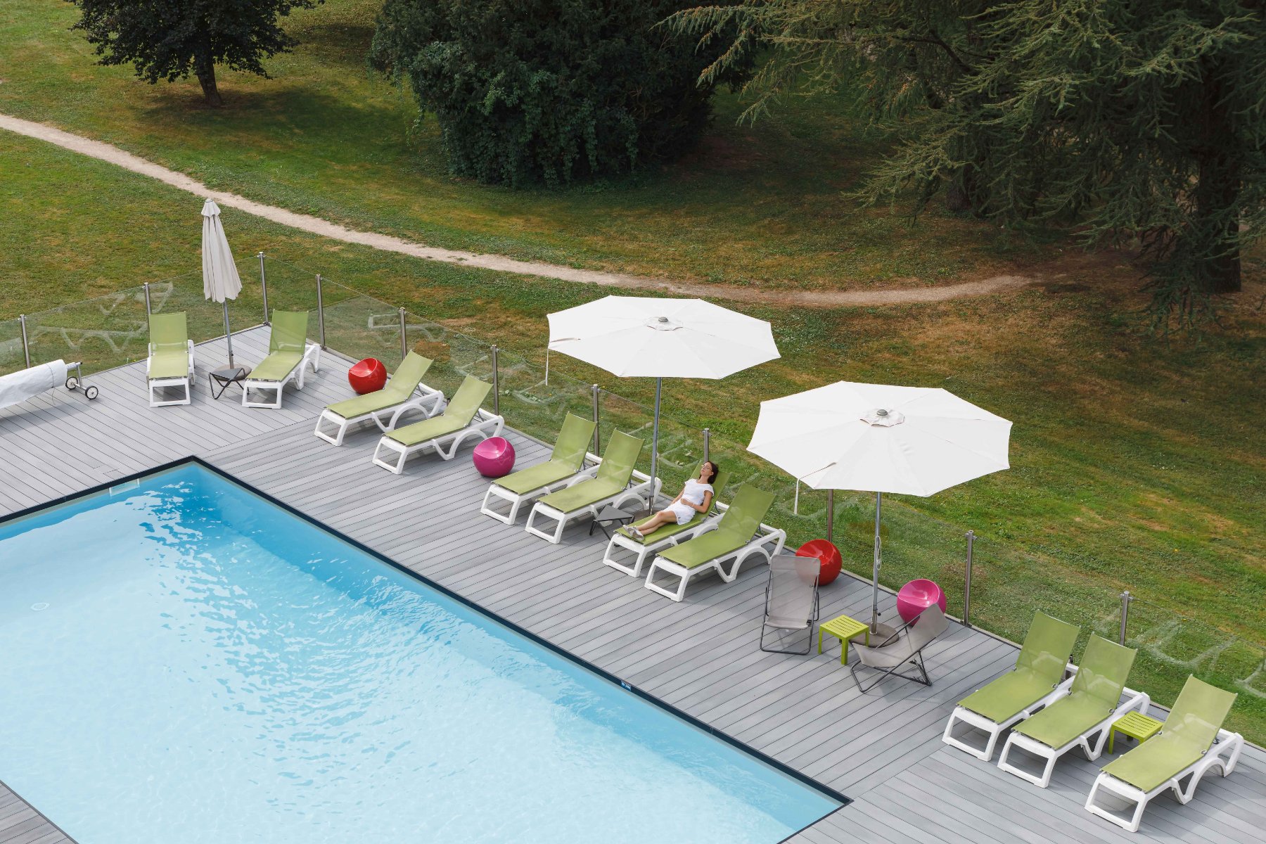 en::Outdoor pool with blue water, wooden deck, sun loungers and parasols, set in a park. fr::Piscine extérieure au fond bleu, terrasse en bois, transats et parasols au cœur d’un parc.