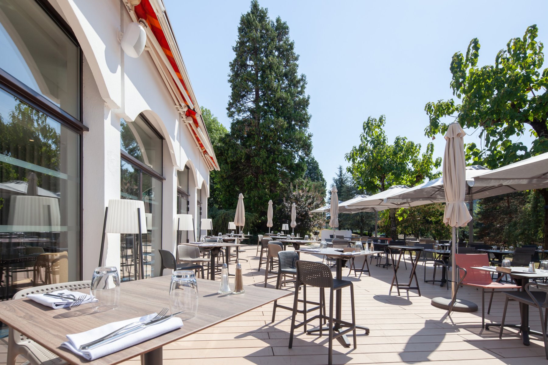 en::Wooden terrace surrounded by trees and pines, with tables and chairs, and open and closed parasols. fr::Terrasse en bois entourée d’arbres et de sapins, avec tables, chaises et parasols ouverts et fermés.