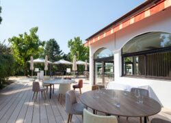 en::Wooden terrace along a tree-lined path, restaurant façade with orange and yellow awnings. fr::Terrasse en bois le long d’une allée arborée, façade du restaurant avec stores orange et jaune.
