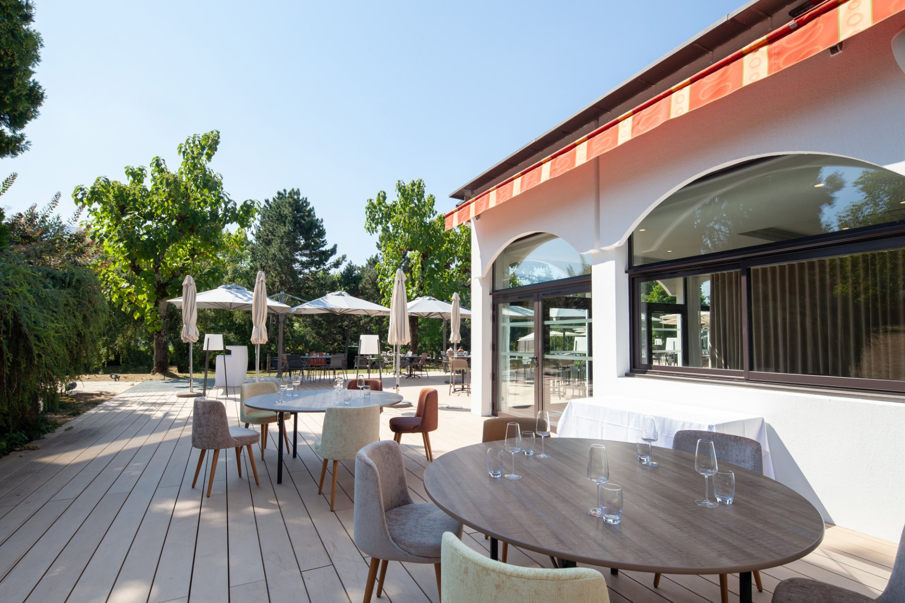 en::Wooden terrace along a tree-lined path, restaurant façade with orange and yellow awnings. fr::Terrasse en bois le long d’une allée arborée, façade du restaurant avec stores orange et jaune.