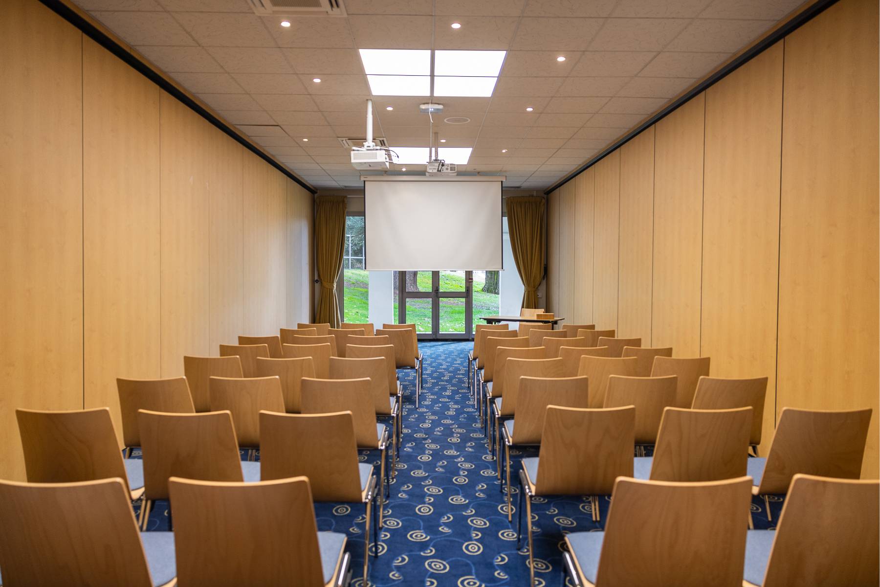 en:: Rows of chairs on both sides of an aisle facing a screen, in front of a glass door with double curtains.fr:: Rangées de chaises de part et d’autre d’une allée face à un écran, devant une porte-fenêtre avec doubles rideaux.