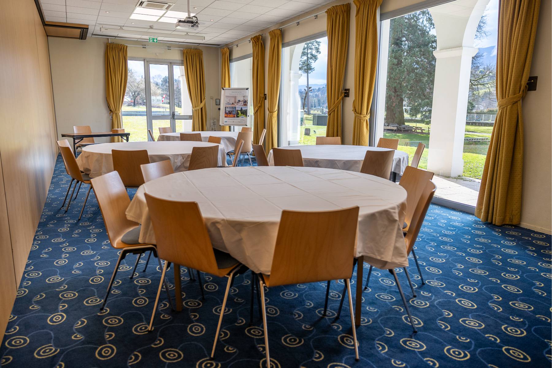 en::Room with glass door and angled windows, blue carpet with orange circles, round tables with tablecloths. fr::Salle avec porte-fenêtre et baies vitrées, moquette bleue à motifs ronds orangés, tables rondes nappées.