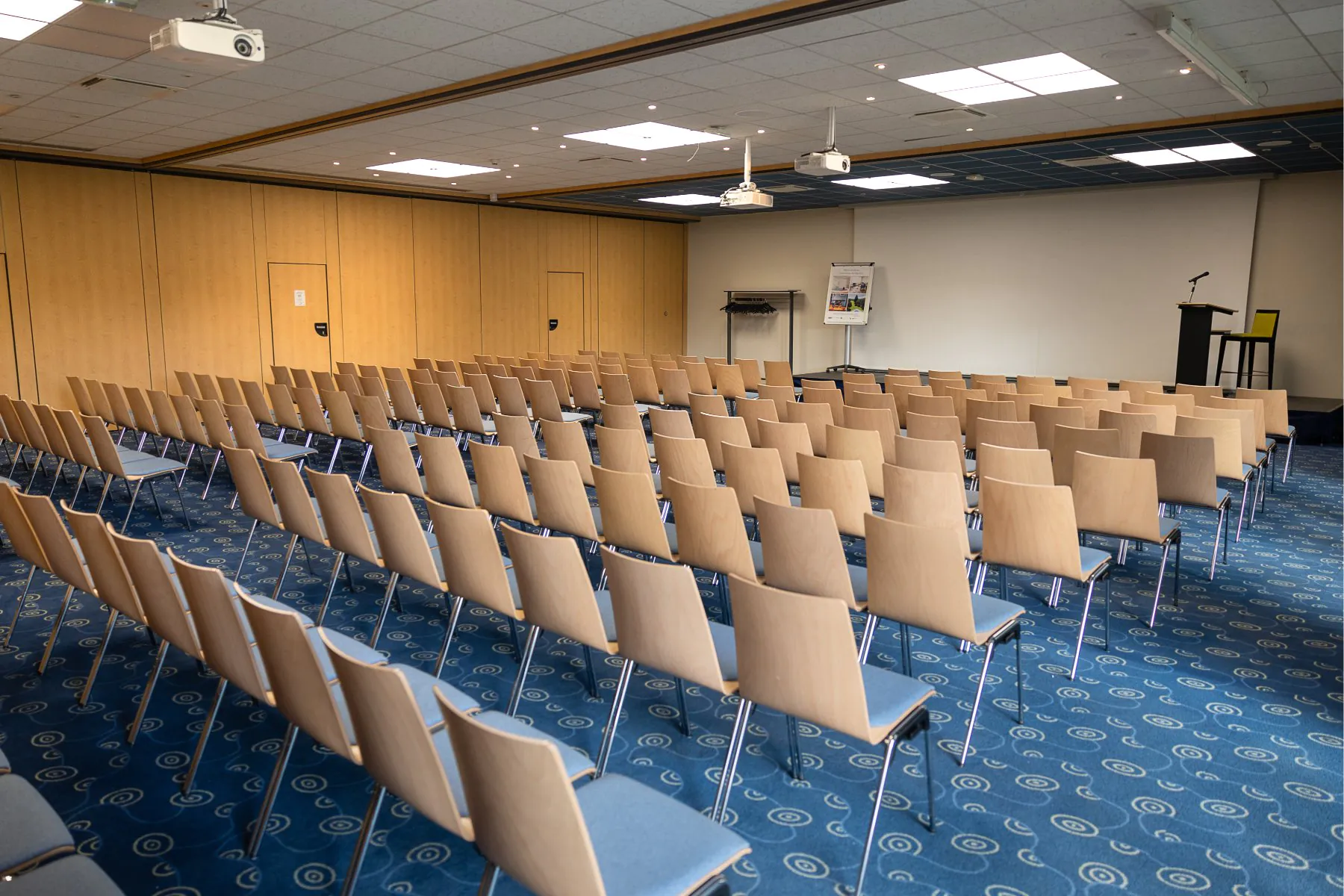 en::Blue chairs in two rows facing a large wall screen, stage with lectern, cloakroom on the left. fr::Chaises bleues en deux rangées face à un grand écran mural, scène avec pupitre, vestiaire à gauche.