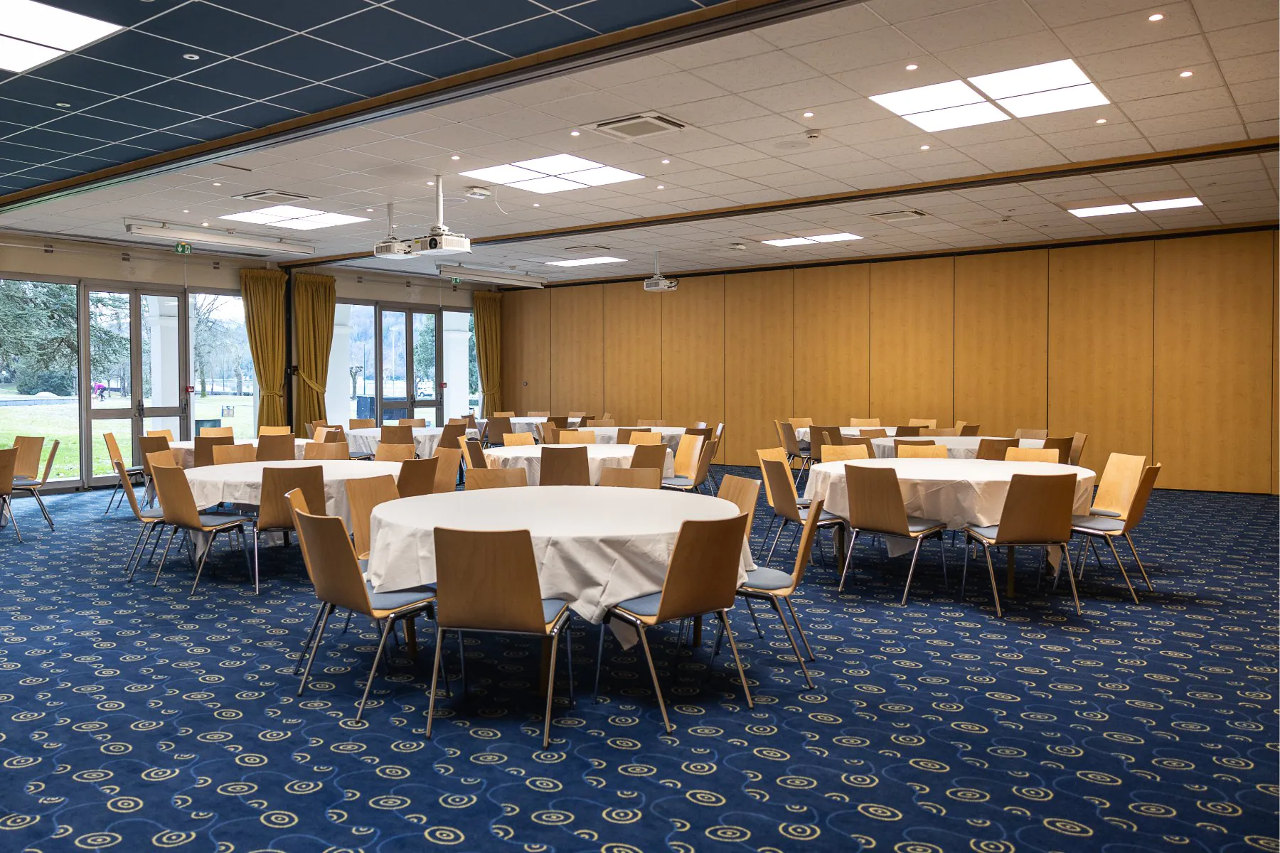 en::Round tables with tablecloths in a room with blue carpet, bay windows overlooking the park, projector equipment. fr::Tables rondes nappées dans une salle avec moquette bleue, baies vitrées sur le parc, équipement vidéoprojecteur.