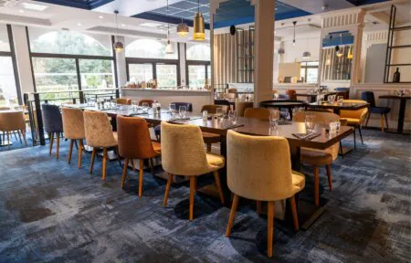 en::Room with blue carpet, blue and white ceiling, square wooden tables, chairs in blue, beige and bronze tones. fr::Salle avec moquette bleue, plafond bleu et blanc, tables carrées en bois, chaises bleues, beiges et bronze.