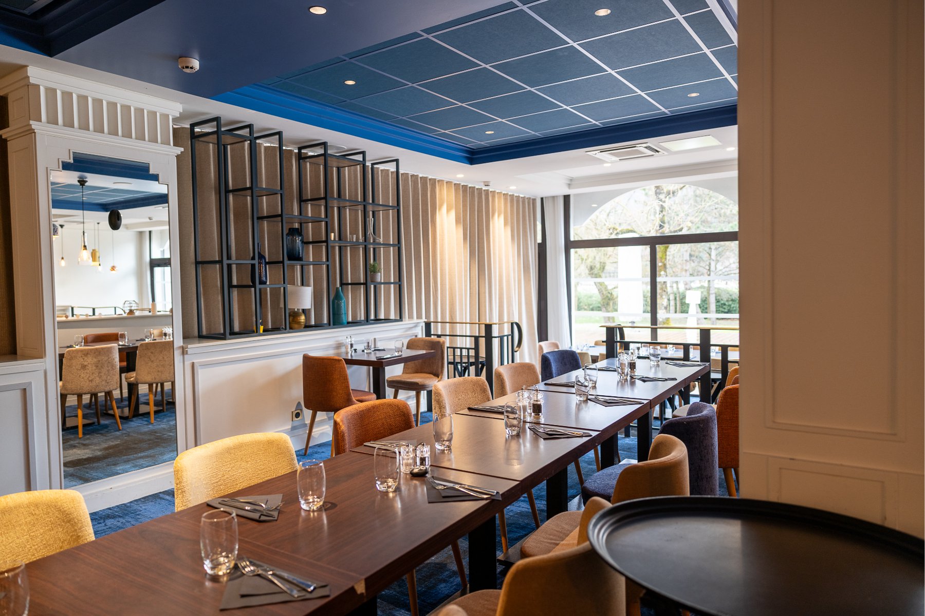 en::Row of square tables aligned across the room, facing a mirrored column defining the group area. fr::Alignement de tables carrées dans la salle, face à une colonne miroir délimitant l’espace groupe.