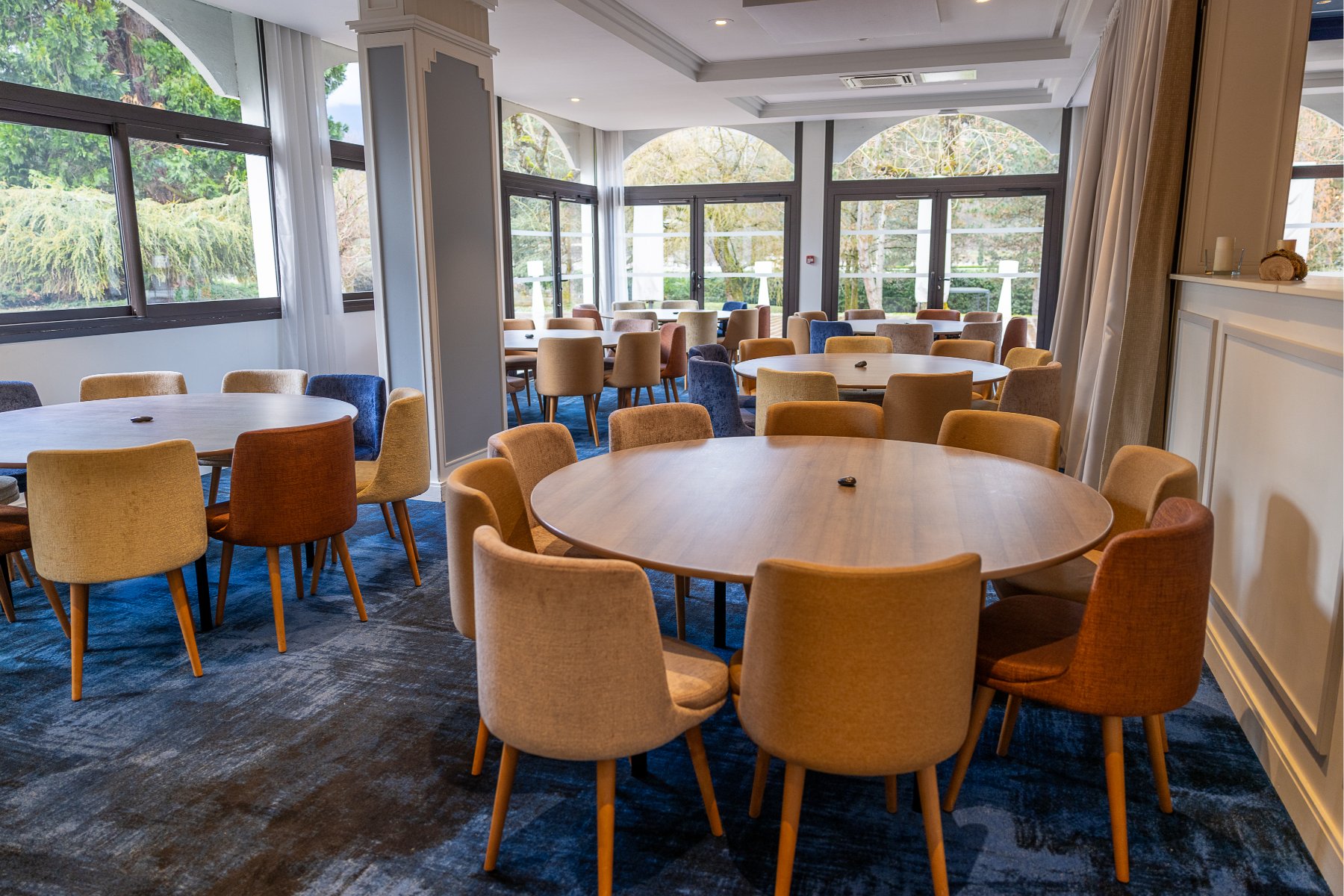en::Private restaurant room with bay windows overlooking a park, featuring large round tables. fr::Salle de restaurant privatisable avec baies vitrées sur parc, composée de grandes tables rondes.