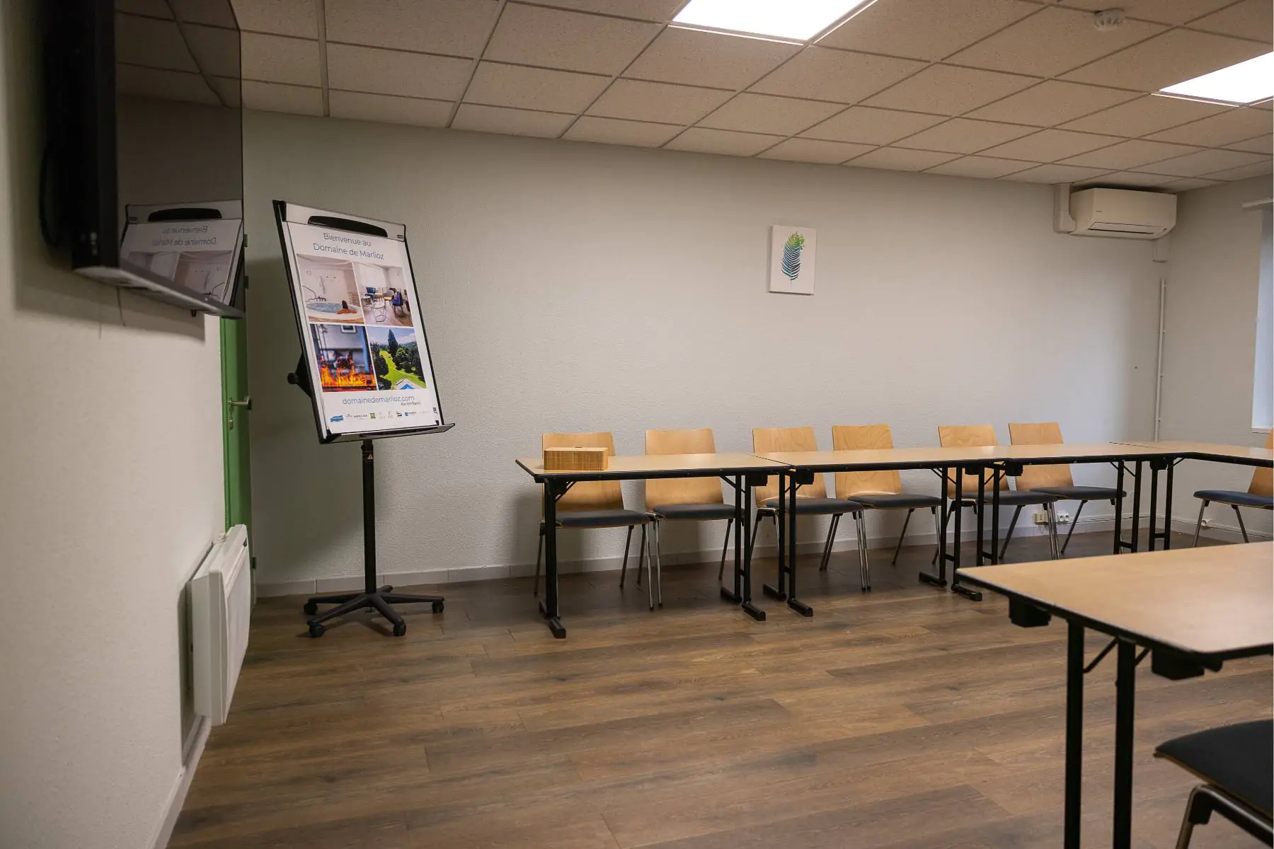en::Rectangular wooden tables arranged in a U-shape, room with heater and air conditioning. fr::Tables rectangulaires en bois en U, salle avec convecteur et climatisation.