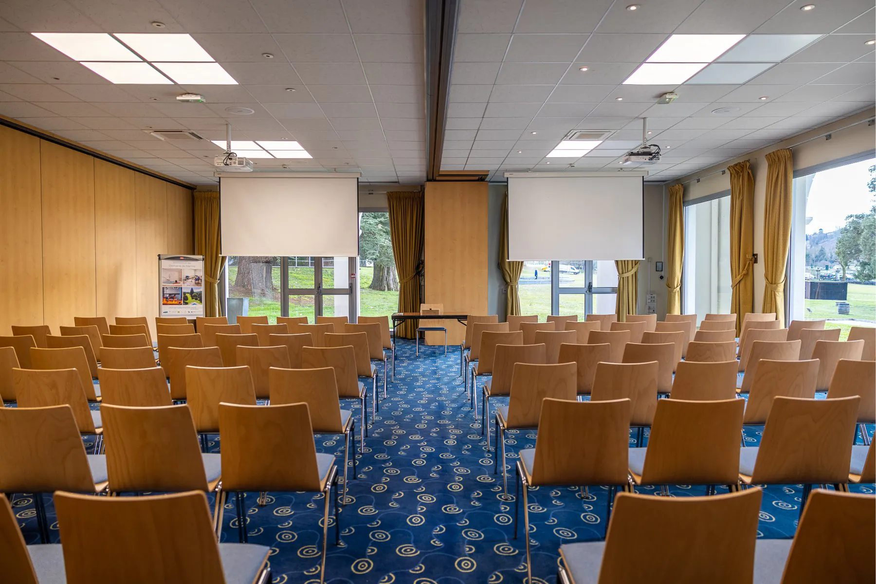 en::Blue chairs in two rows facing two ceiling-mounted screens, partially covering glass doors to the park. fr::Chaises bleues en deux rangées face à deux écrans descendus du plafond, occultant partiellement les portes-fenêtres vers le parc.