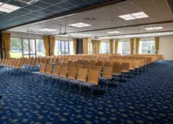 en::Large meeting room with many chairs facing a stage, two walls with bay windows. fr::Grande salle de réunion avec nombreuses chaises face à une estrade, deux murs en baies vitrées.