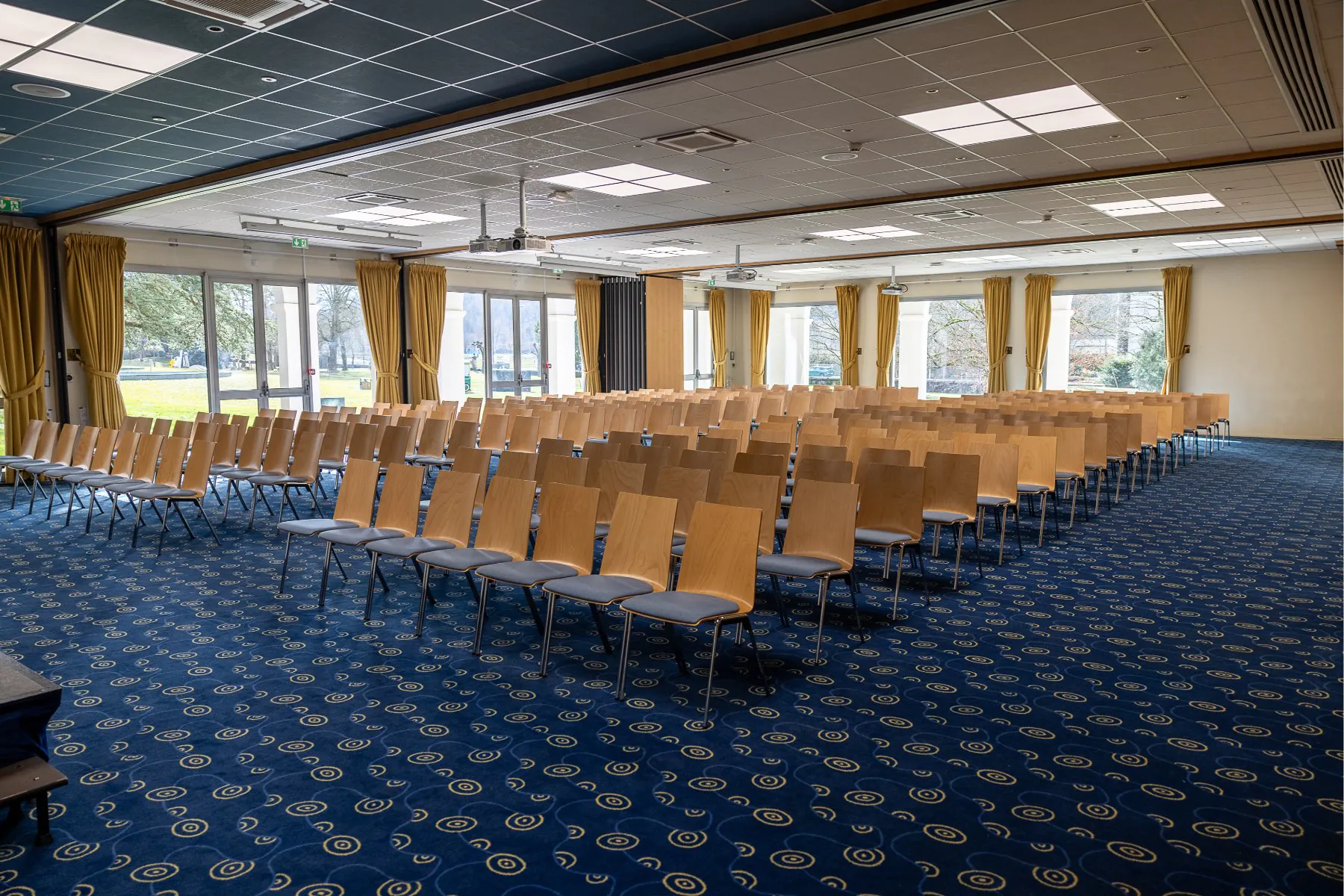 en::Large meeting room with many chairs facing a stage, two walls with bay windows. fr::Grande salle de réunion avec nombreuses chaises face à une estrade, deux murs en baies vitrées.