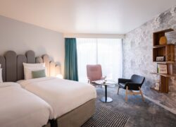 en::Twin beds with grey headboards facing a wall with grey cloud-patterned wallpaper, decorative shelves, pastel-toned lounge area. fr::Lits twin avec têtes de lit grises face à un mur à motifs nuageux gris, étagères décoratives, coin salon aux tons pastel.