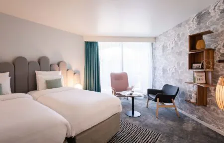 en::Twin beds with grey headboards facing a wall with grey cloud-patterned wallpaper, decorative shelves, pastel-toned lounge area. fr::Lits twin avec têtes de lit grises face à un mur à motifs nuageux gris, étagères décoratives, coin salon aux tons pastel.