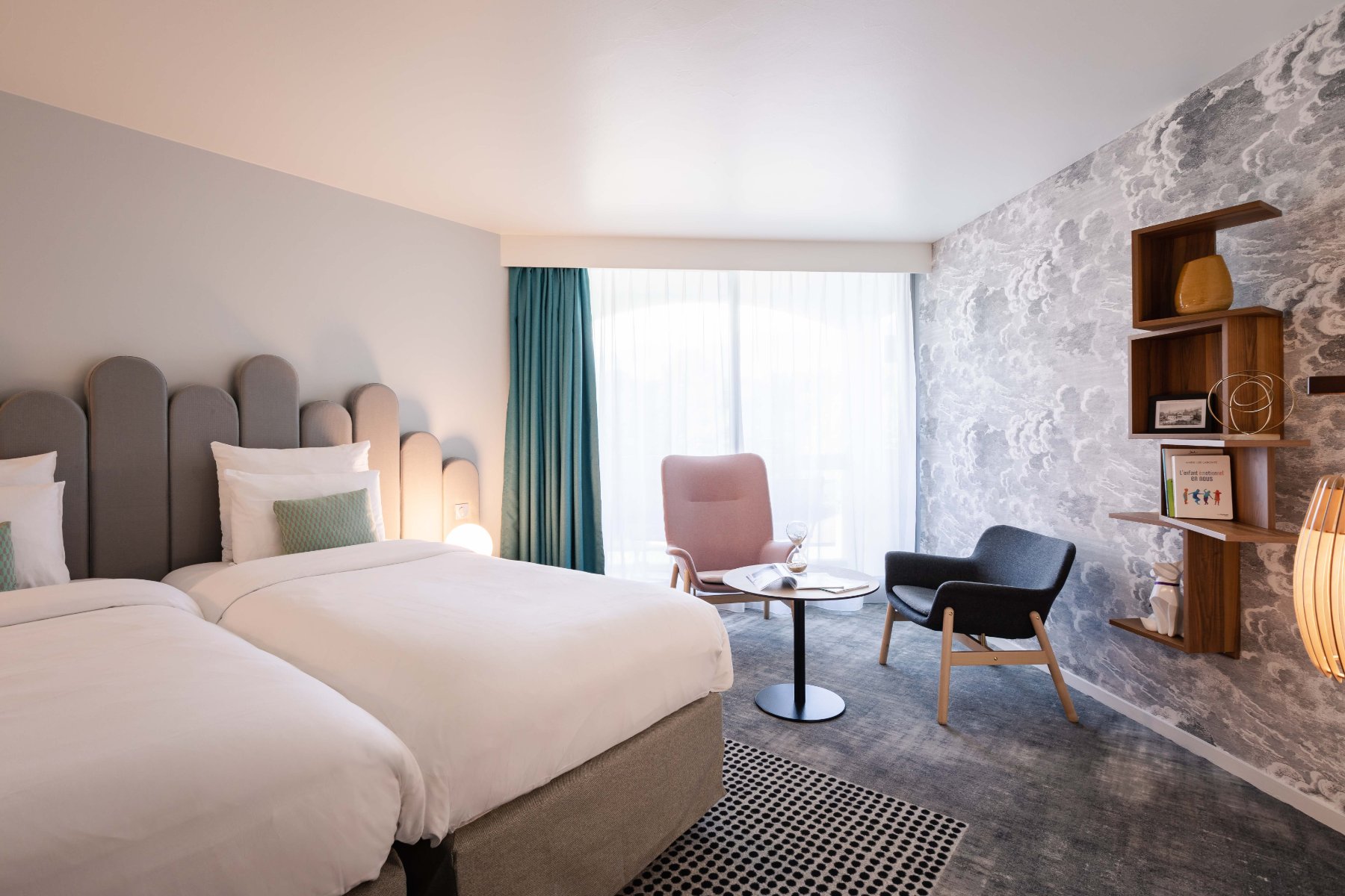 en::Twin beds with grey headboards facing a wall with grey cloud-patterned wallpaper, decorative shelves, pastel-toned lounge area. fr::Lits twin avec têtes de lit grises face à un mur à motifs nuageux gris, étagères décoratives, coin salon aux tons pastel.