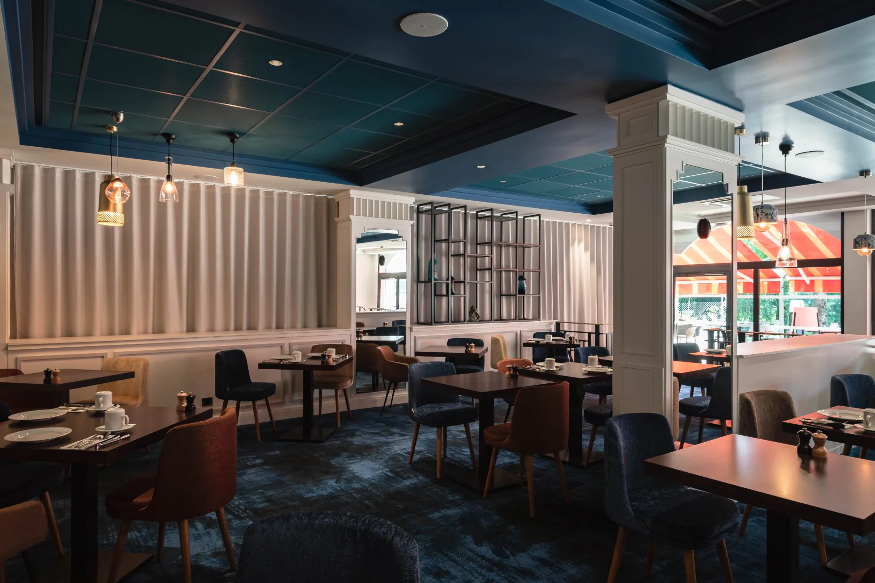en::Restaurant room with royal blue ceiling and carpet, white walls and sheer curtains, mirrored columns, tables and chairs with terrace view. fr::Salle de restaurant avec plafond et moquette bleu roi, murs et voilages blancs, colonnes miroir, tables et chaises avec vue sur la terrasse.