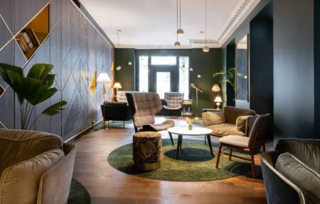 en::Armchairs and sofas in various styles and shapes arranged around coffee tables, with rugs on the floor and the entrance door at the back of the living area. fr::Fauteuils et canapés de styles et formes variés autour de tables basses, tapis au sol, et porte d’entrée au fond de la pièce à vivre.