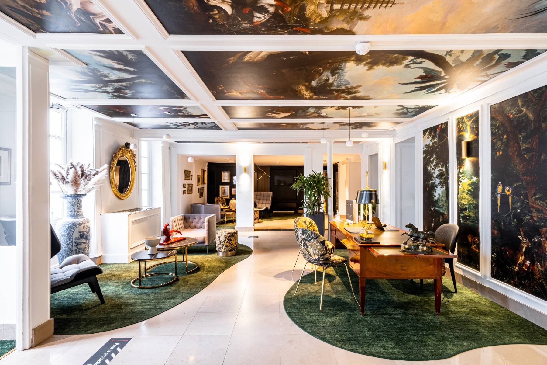 en::Hotel reception area with decorative tapestry on the ceiling and walls, depicting landscapes and exotic animals such as parrots, geese, and horses. fr::Réception de l’hôtel avec tapisserie décorative au plafond et sur les murs, représentant des paysages et animaux exotiques comme des perroquets, oies et chevaux.