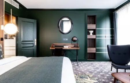 en::Large double bed set against a wall combining wood and brass, wooden secretary desk with an oval mirror above, set against a green-painted wall. fr::Grand lit double contre un mur habillé de bois et de laiton, bureau secrétaire en bois avec miroir ovale au-dessus, contre un mur peint en vert.