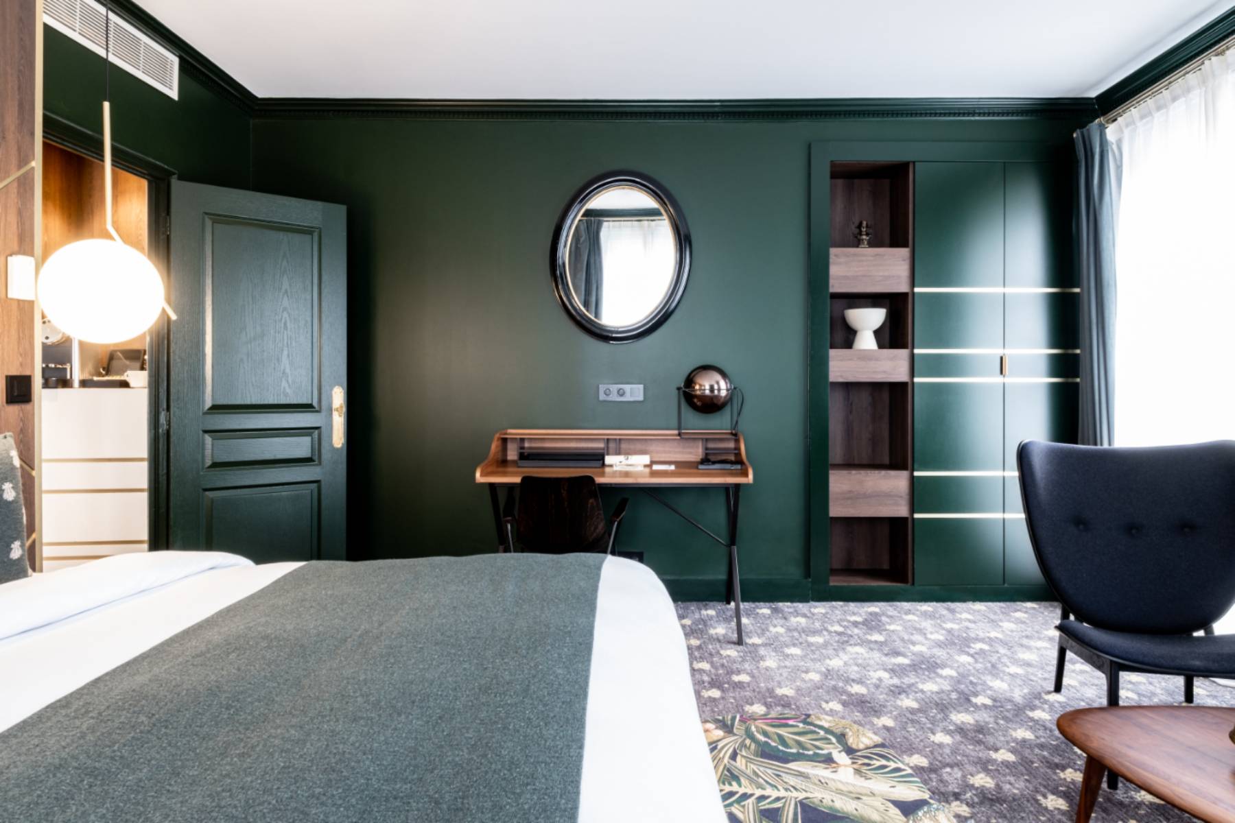 en::Large double bed set against a wall combining wood and brass, wooden secretary desk with an oval mirror above, set against a green-painted wall. fr::Grand lit double contre un mur habillé de bois et de laiton, bureau secrétaire en bois avec miroir ovale au-dessus, contre un mur peint en vert.