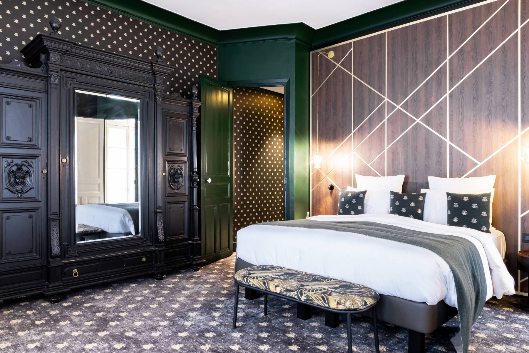 en::Large double bed set against a wall combining wood and brass, antique black-painted wooden wardrobe with a mirror, carpeted floor, and wallpaper featuring the hotel’s signature pattern. fr::Grand lit double contre un mur habillé de bois et de laiton, armoire ancienne en bois peint en noir avec miroir, moquette et tapisserie aux motifs signature de l’hôtel.
