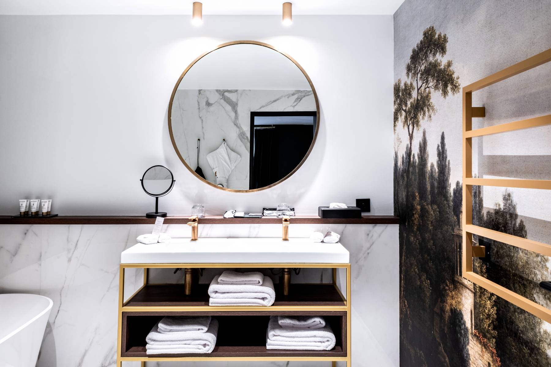 en::Bathroom with a designer bathtub set against a marble wall, rectangular sink with brass and wood shelving, a round gold mirror, and decorative wall tapestry. fr::Salle de bain avec baignoire design adossée à un mur en marbre, lavabo rectangulaire avec étagère en laiton et bois, miroir rond doré et tapisserie murale décorative.