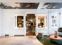 en::Bar entrance framed by white columns, ceiling decorated with landscapes and animals, fireplace in the background, long counter on the left, and a cosy library lounge on the right. fr::Entrée du bar entre colonnes blanches, plafond décoré de paysages et animaux, cheminée en fond, comptoir sur toute la longueur à gauche et salon bibliothèque cosy à droite.