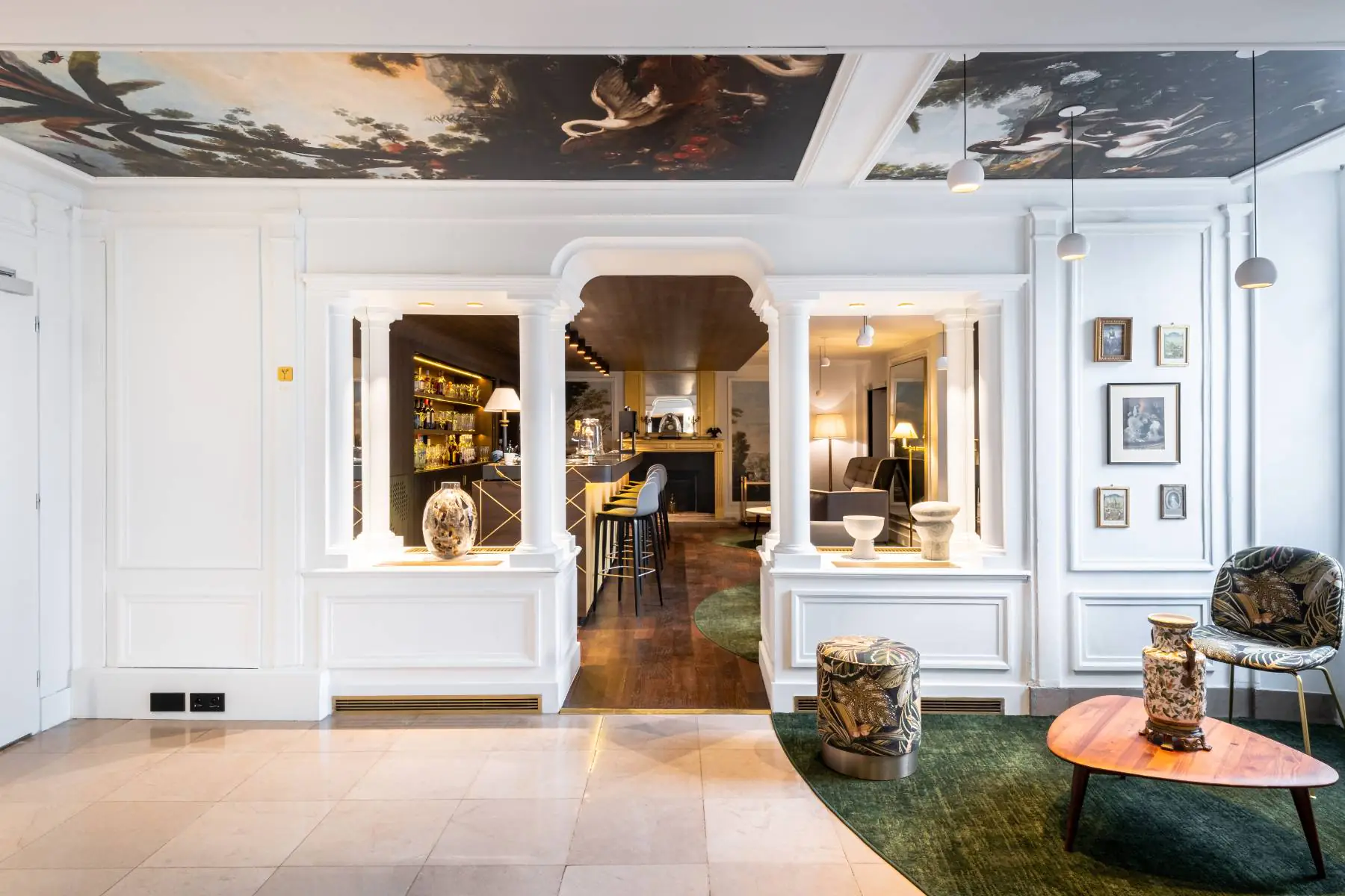 en::Bar entrance framed by white columns, ceiling decorated with landscapes and animals, fireplace in the background, long counter on the left, and a cosy library lounge on the right. fr::Entrée du bar entre colonnes blanches, plafond décoré de paysages et animaux, cheminée en fond, comptoir sur toute la longueur à gauche et salon bibliothèque cosy à droite.