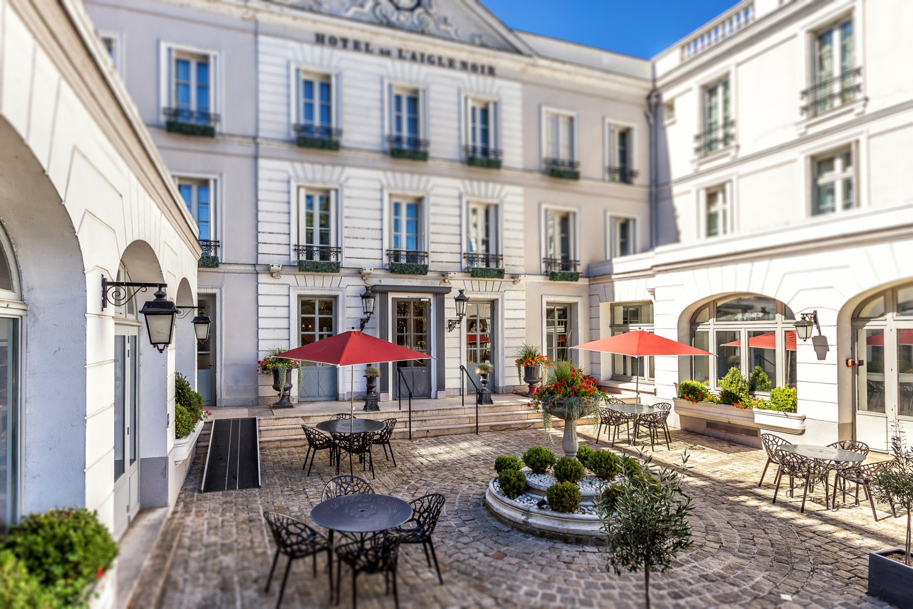en::Hotel inner courtyard with central greenery, black metal round tables and matching chairs, red parasols, and U-shaped buildings surrounding the space. fr::Cour intérieure de l’hôtel avec végétation centrale, tables rondes en métal noir et chaises assorties, parasols rouges, et bâtiments en U entourant l’espace.
