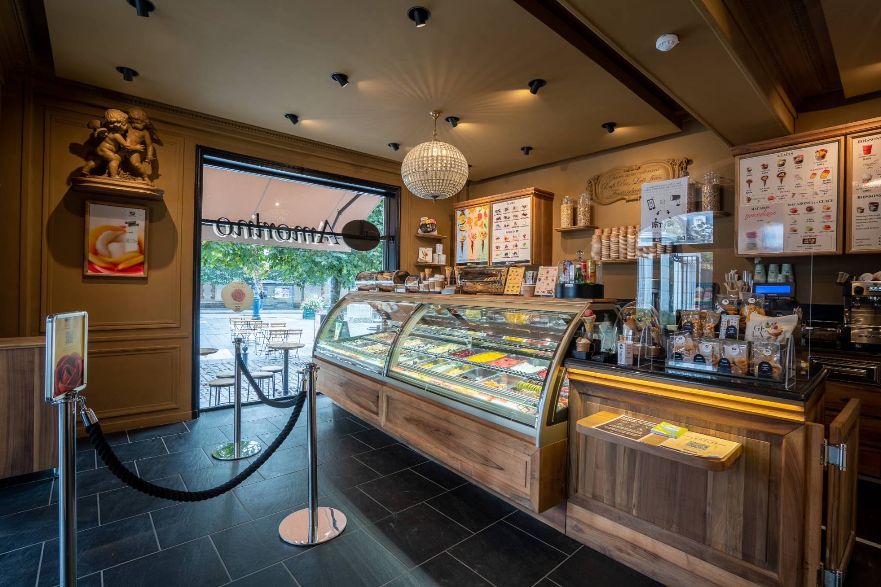 en::Amorino wooden counter offering a wide selection of ice creams, decorative corner statues, and a view of the terrace through the display window. fr::Comptoir Amorino en bois proposant un large choix de glaces, statues décoratives en angle, et vue sur la terrasse depuis la vitrine.