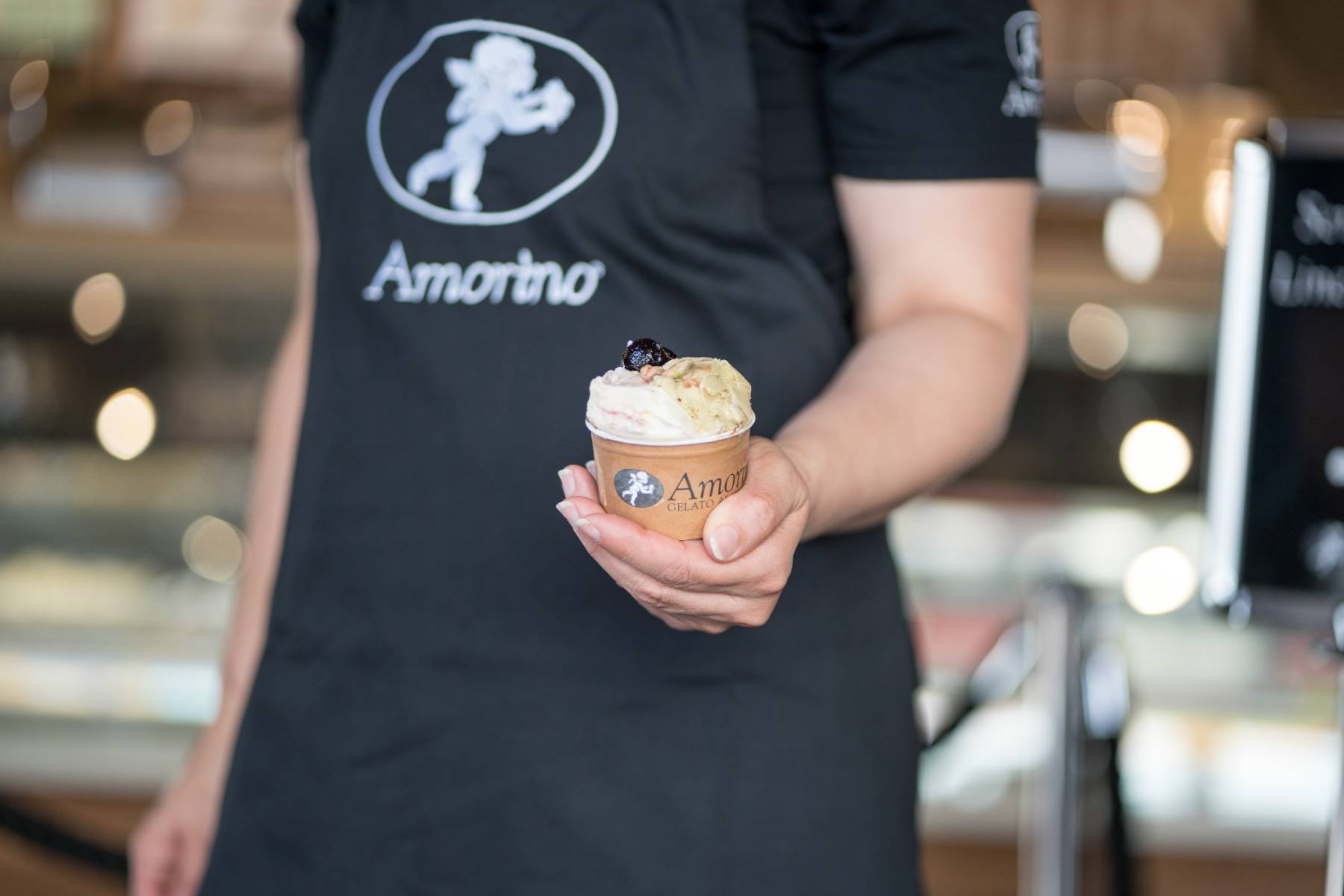 en::Person wearing an Amorino apron handing over a two-flavour ice cream topped with a cherry. fr::Personne en tablier Amorino tendant un pot de glace à deux parfums surmonté d’une cerise.