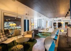 en::Bar counter with six bar stools, large armchairs and coffee tables opposite, and a large wall mirror reflecting the back of the bar. fr::Comptoir de bar avec six chaises hautes, larges fauteuils et tables basses en face, et grand miroir mural reflétant l’arrière du bar.