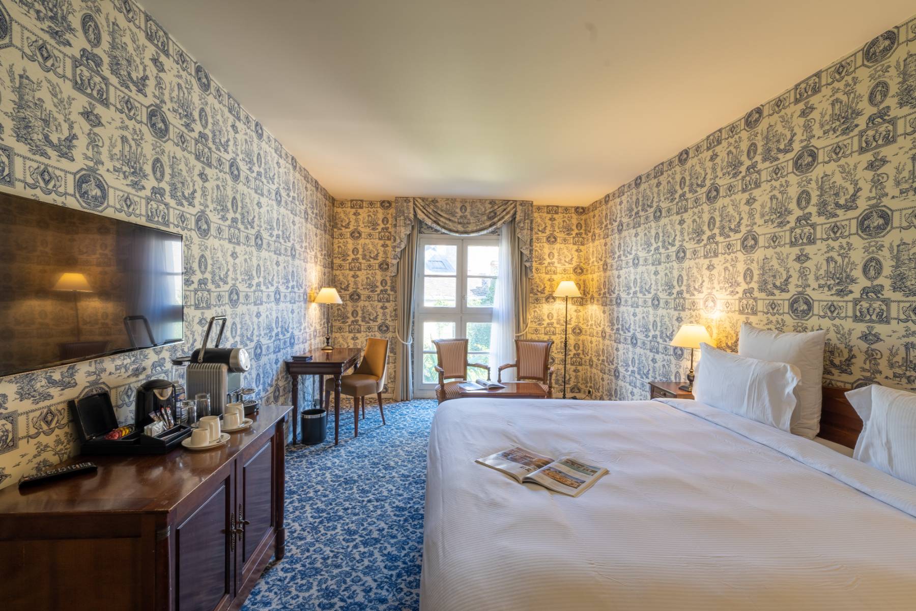 en::Second Empire-style room in white and blue tones, large window with views over the city and greenery, lamps switched on. fr::Chambre Second Empire aux tons blancs et bleus, grande fenêtre avec vue sur la ville et la végétation, lampes allumées.