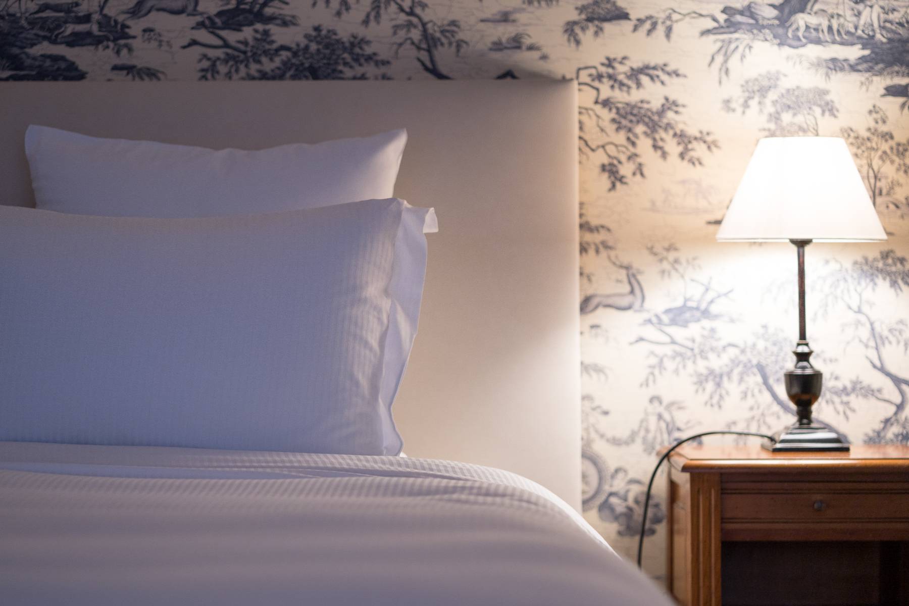 en::Bed with headboard, white pillows and duvet, blue and white forest-patterned wallpaper, and a wooden bedside table. fr::Lit avec tête de lit, oreillers et couette blancs, tapisserie aux motifs forestiers bleus et blancs, et table de chevet en bois.