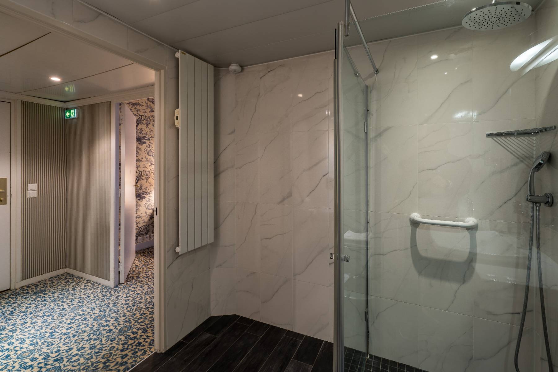 en::Accessible walk-in shower room, open door to the corridor with blue and white patterned carpet and wallpaper. fr::Salle de douche PMR à l’italienne, porte ouverte sur le couloir avec moquette et tapisserie aux motifs bleus et blancs.