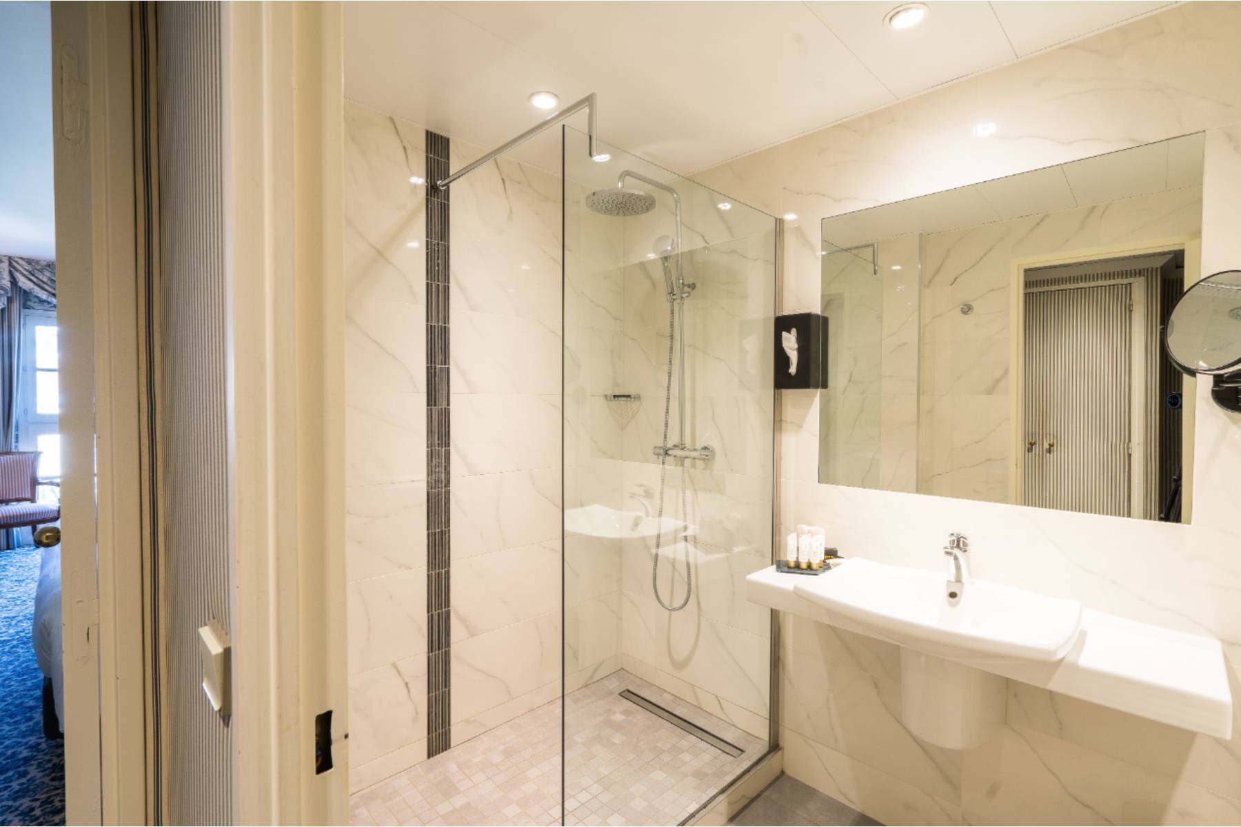 en::Walk-in shower room with a white sink, open door to the bedroom with blue and white patterned carpet, thick curtains, and a wooden chair with white and burgundy striped fabric. fr::Salle de douche à l’italienne avec lavabo blanc, porte ouverte sur la chambre avec moquette imprimée bleu et blanc, rideaux épais et chaise en bois au tissu rayé blanc et bordeaux.