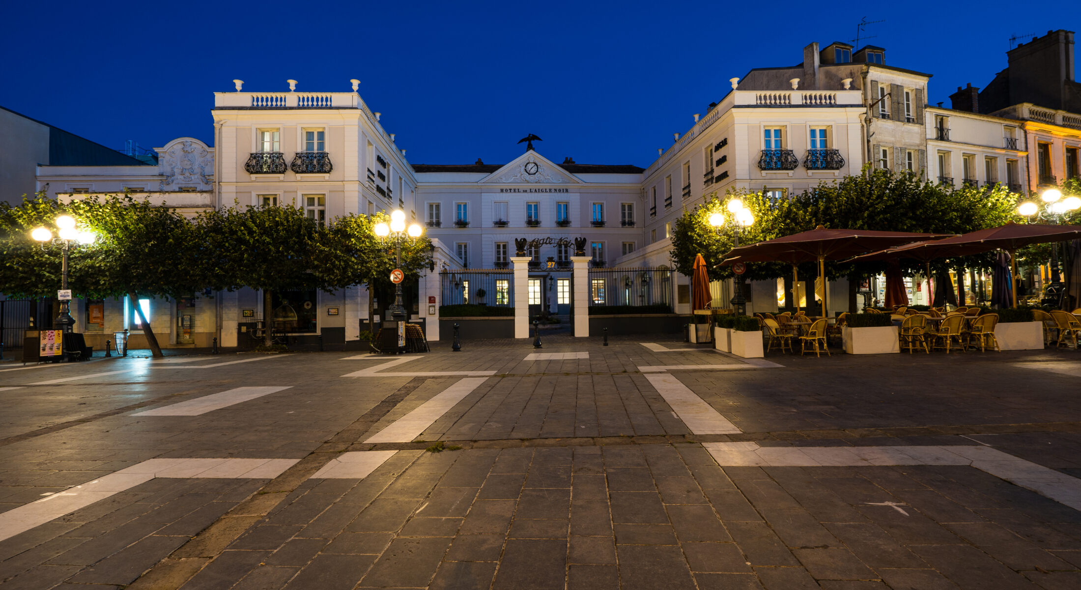 en::U-shaped building seen at night from a square lit by street lamps, lined with trees on either side of the gate and entrance porch. fr::Bâtiment en U vu de nuit depuis une place éclairée par des lampadaires, bordée d’arbres de part et d’autre de la grille et du porche d’entrée.